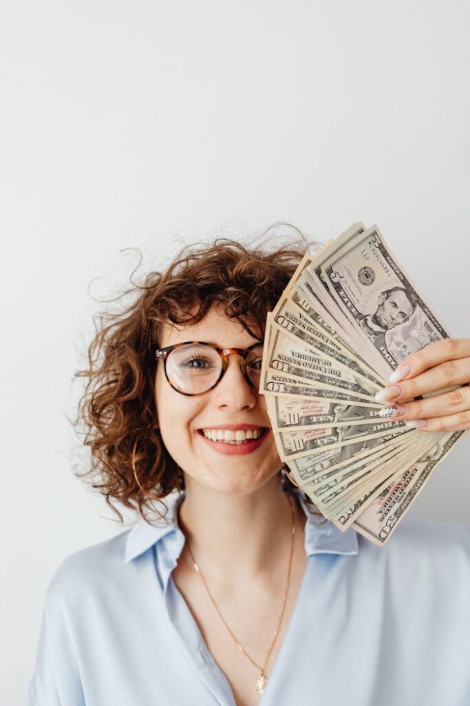 Cheerful woman with curly hair and glasses holding a fan of US dollar bills.